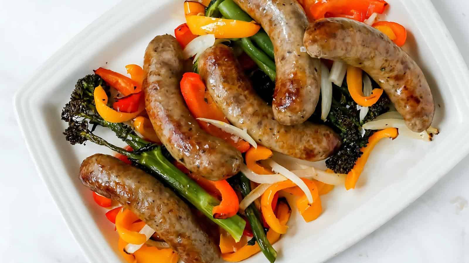 Plate with cooked sausages on top of mixed vegetables, including bell peppers, onions, and broccolini.