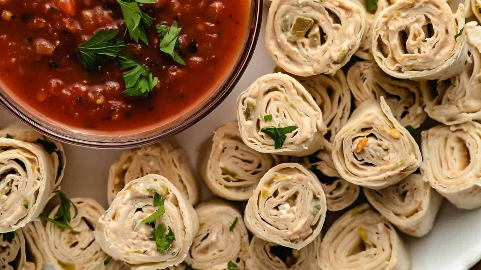 Close-up of tortilla pinwheels with a creamy filling, garnished with herbs, next to a bowl of red salsa topped with cilantro.
