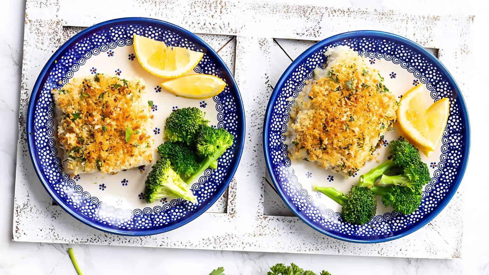 Two blue plates with breaded fish fillets, broccoli, and lemon wedges on a white tray.