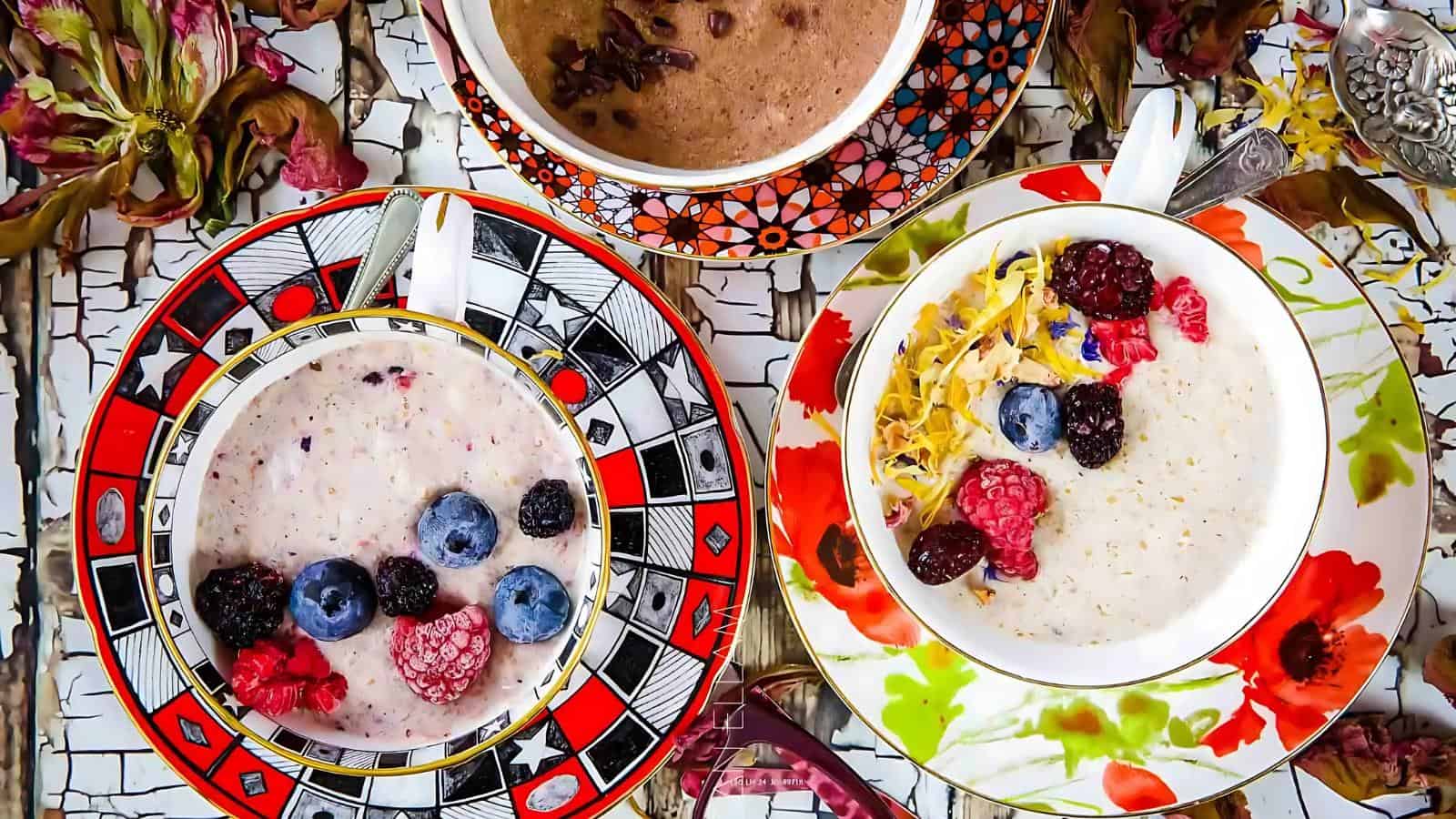 Three teacups with patterns contain oatmeal topped with assorted berries and edible flowers, displayed on a wooden surface with scattered dried leaves.