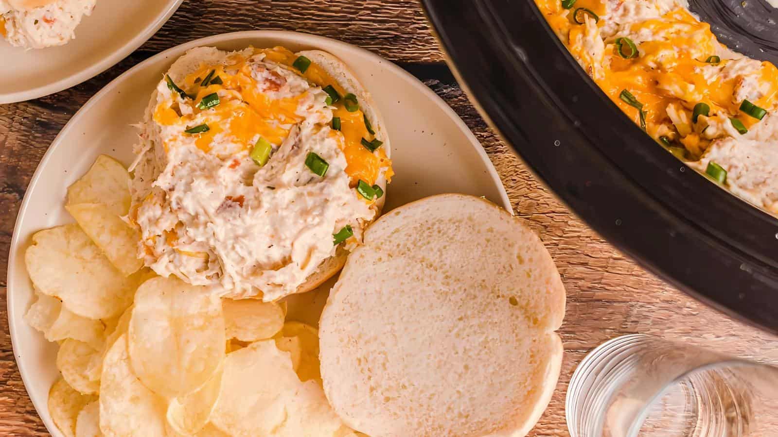 Plate with a sandwich topped with shredded cheese and chives, served with potato chips. Nearby, an empty bun, a glass of water, and a partial view of a crockpot are visible.