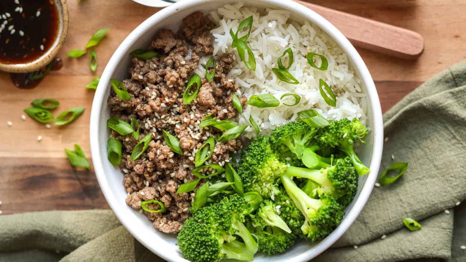 A bowl with white rice, seasoned ground beef, and steamed broccoli, garnished with sliced green onions and sesame seeds. A small dish of sauce is visible in the background.