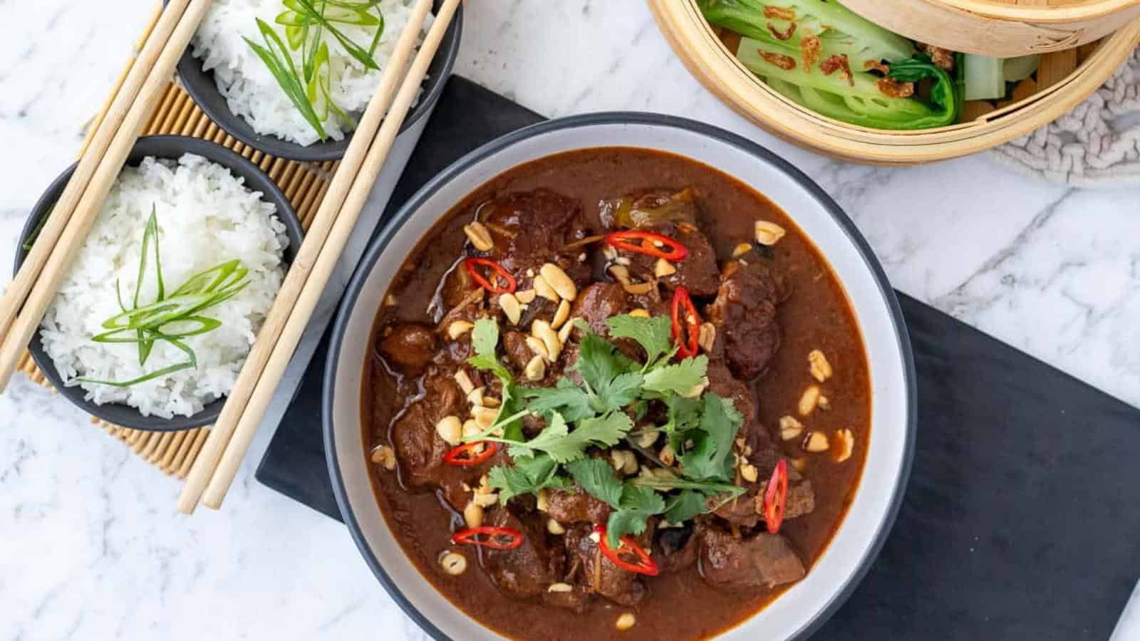 A bowl of rich stew garnished with herbs and sliced chilies, accompanied by two bowls of rice with chopsticks and a bamboo steamer of greens on a marble surface.