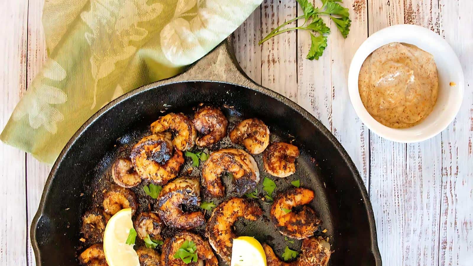 A skillet of seasoned, cooked shrimp garnished with lemon slices and herbs, accompanied by a small bowl of sauce on the side.