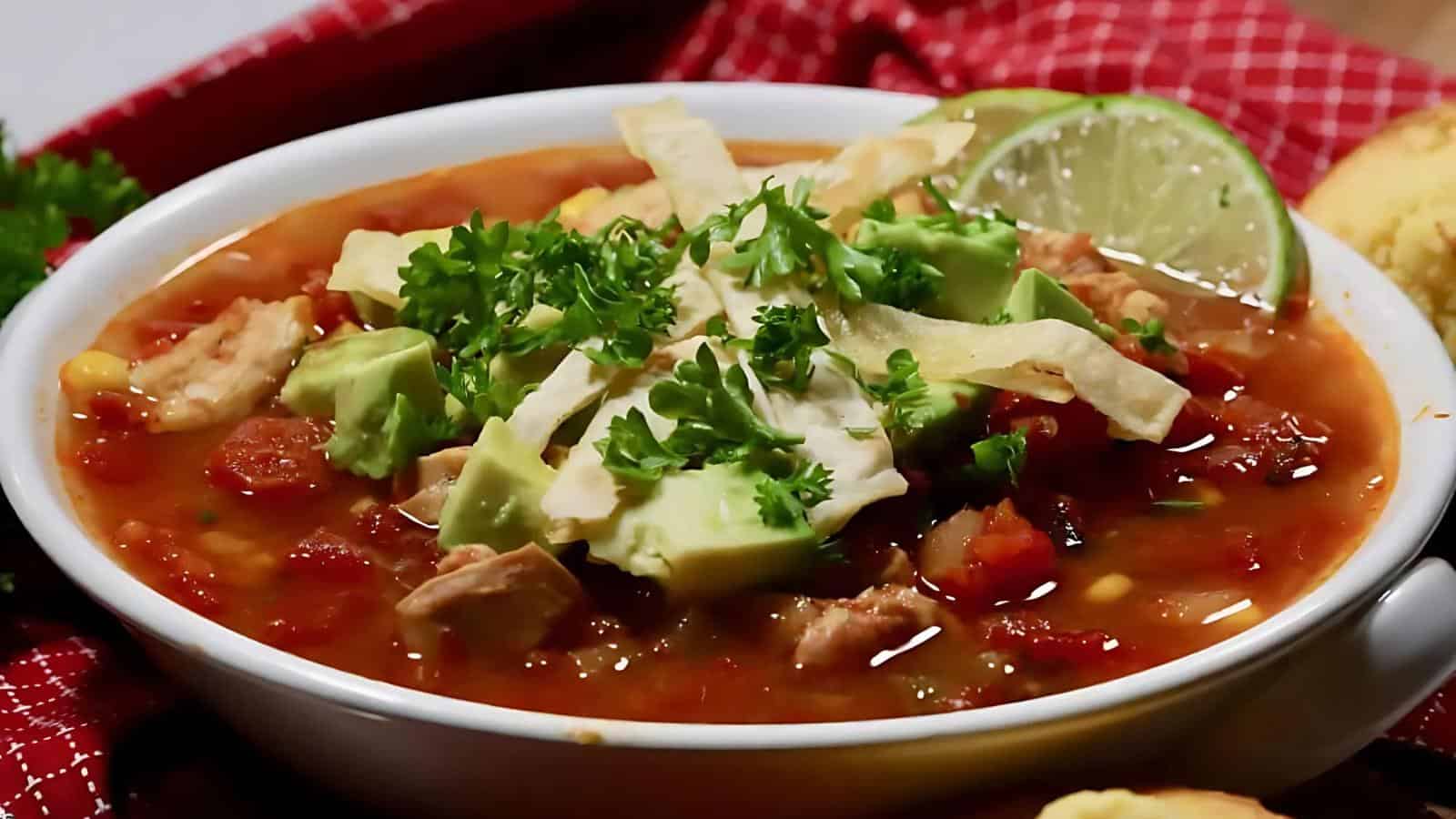 A bowl of chicken tortilla soup garnished with avocado, tortilla strips, parsley, and lime. Red napkin partially visible in the background.