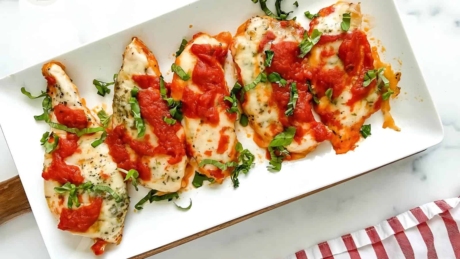 Grilled chicken breasts topped with melted cheese, tomato sauce, and fresh basil on a white rectangular plate.