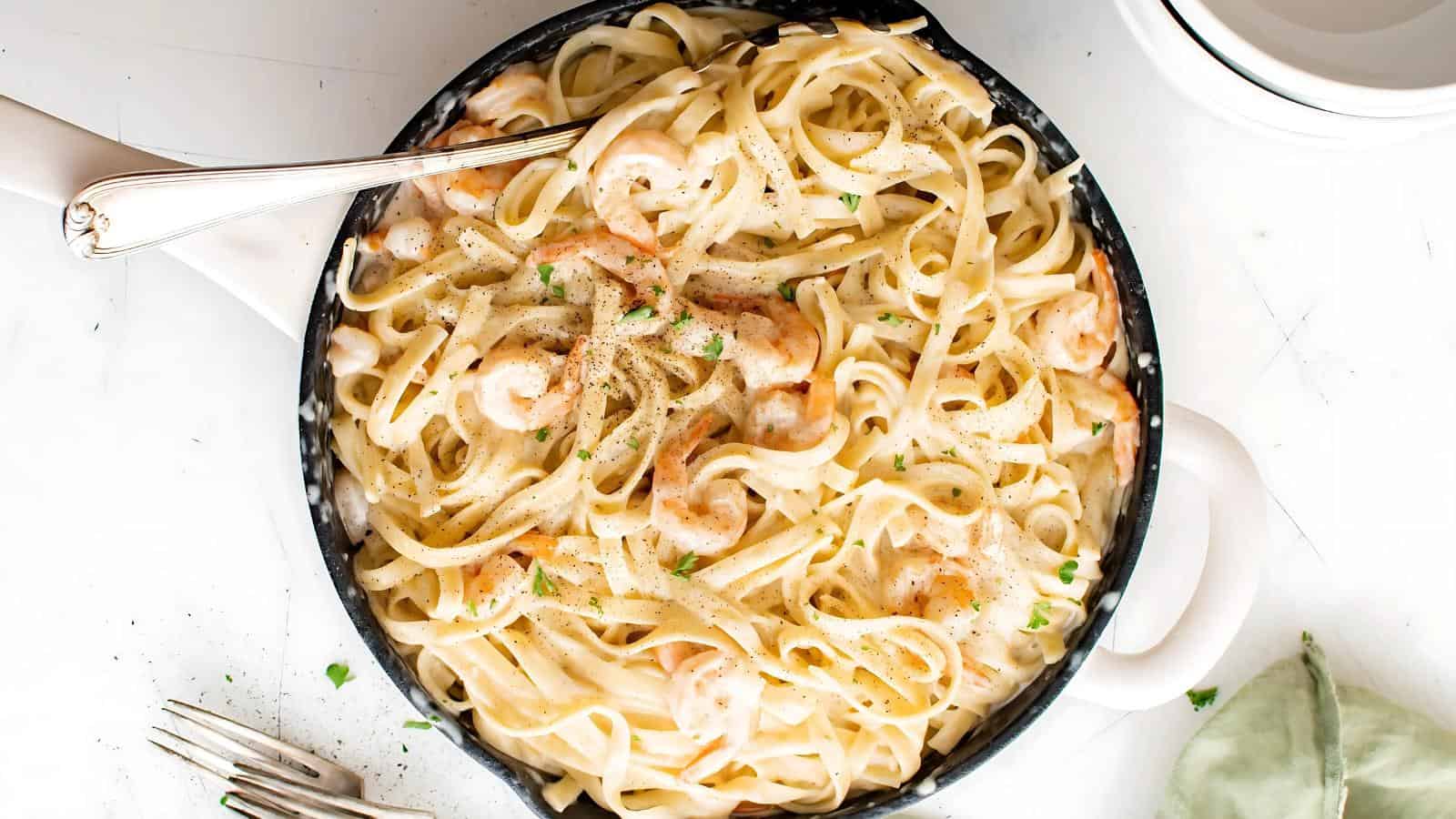 A pan of creamy shrimp fettuccine garnished with herbs, with a silver fork resting on the side.