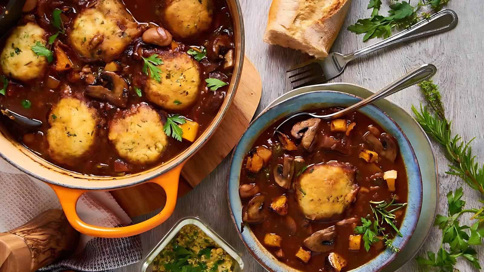 Stew with dumplings in an orange pot and a blue bowl, garnished with parsley. A piece of bread, a fork, and herbs are on a light wooden surface.