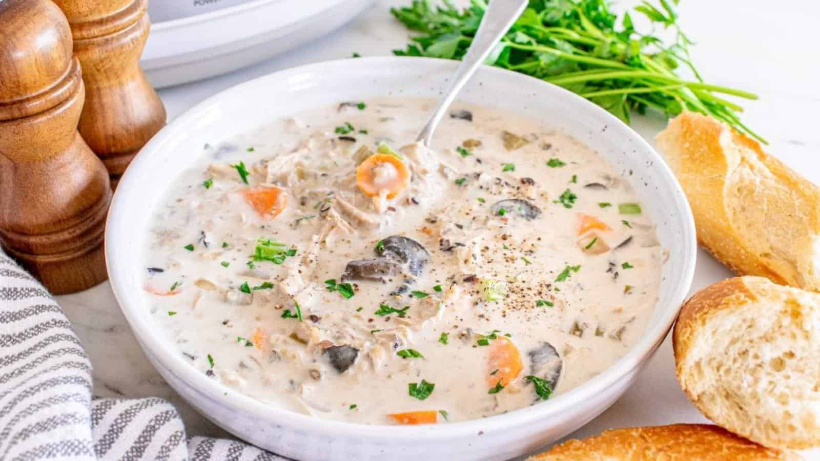 A bowl of creamy chicken and mushroom soup garnished with chopped parsley, served with slices of crusty bread. Pepper mills and fresh herbs are visible in the background.