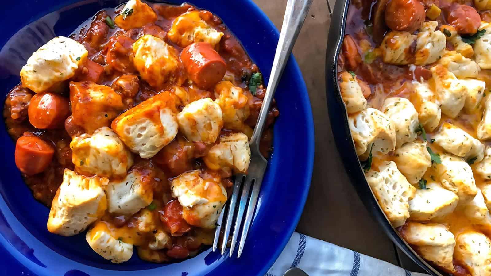A blue plate filled with biscuit pieces, sliced sausages, and a tomato-based sauce; next to a skillet with the same dish.
