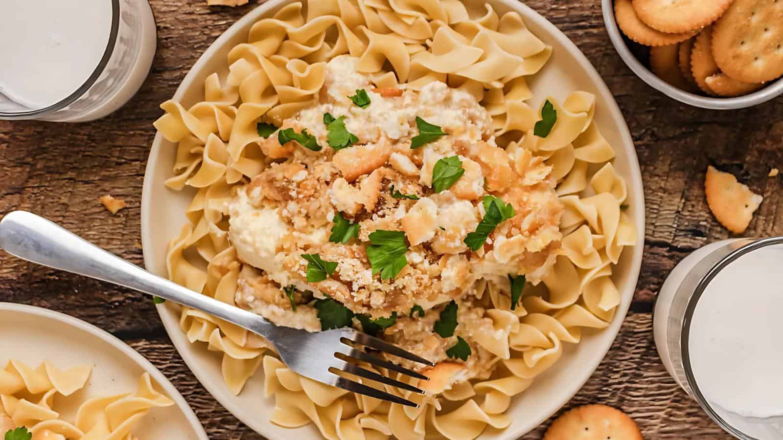 A plate of pasta topped with a creamy sauce and garnished with chopped parsley. A fork rests on the plate. Two glasses of water and a bowl of crackers are nearby.