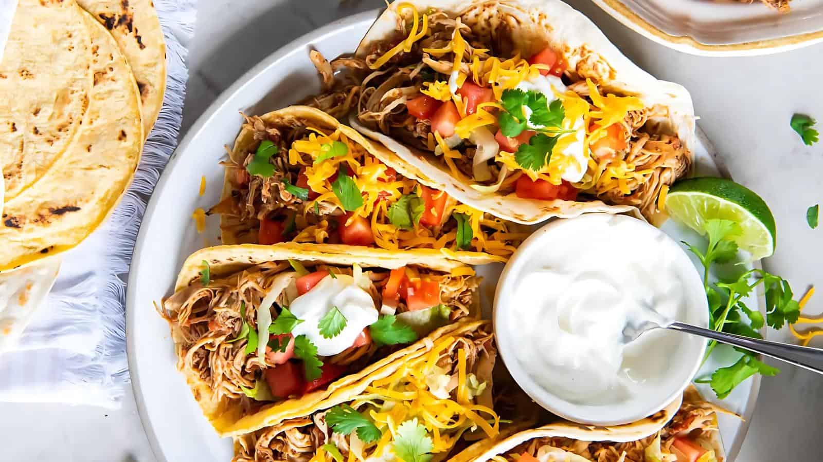 Three tacos filled with shredded chicken, diced tomatoes, cheese, sour cream, and cilantro on a plate with a side of sour cream and a lime wedge.