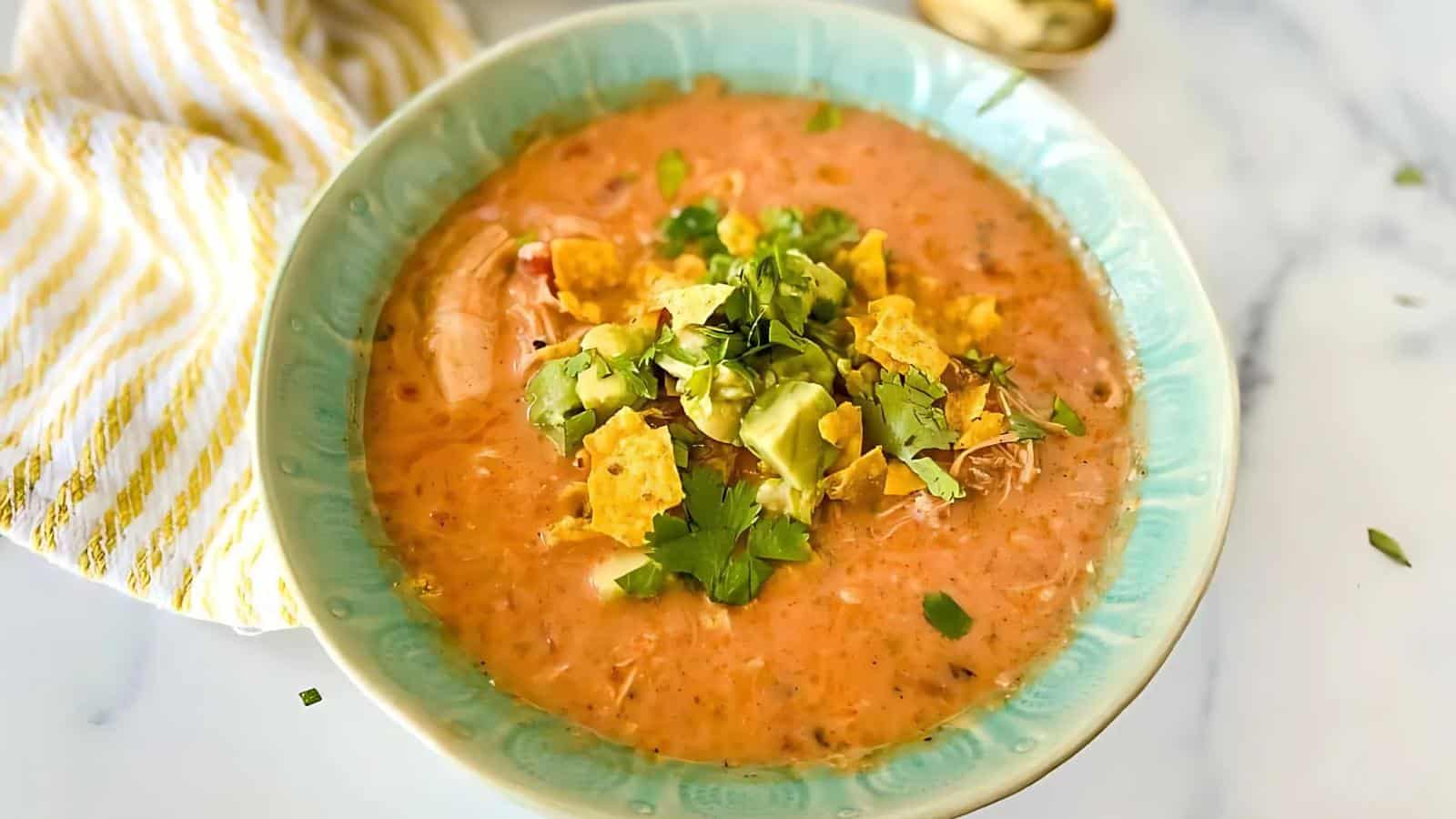 A bowl of creamy soup topped with crushed chips, avocado, and chopped herbs, served in a light blue bowl on a marble surface. A striped napkin and spoon are nearby.