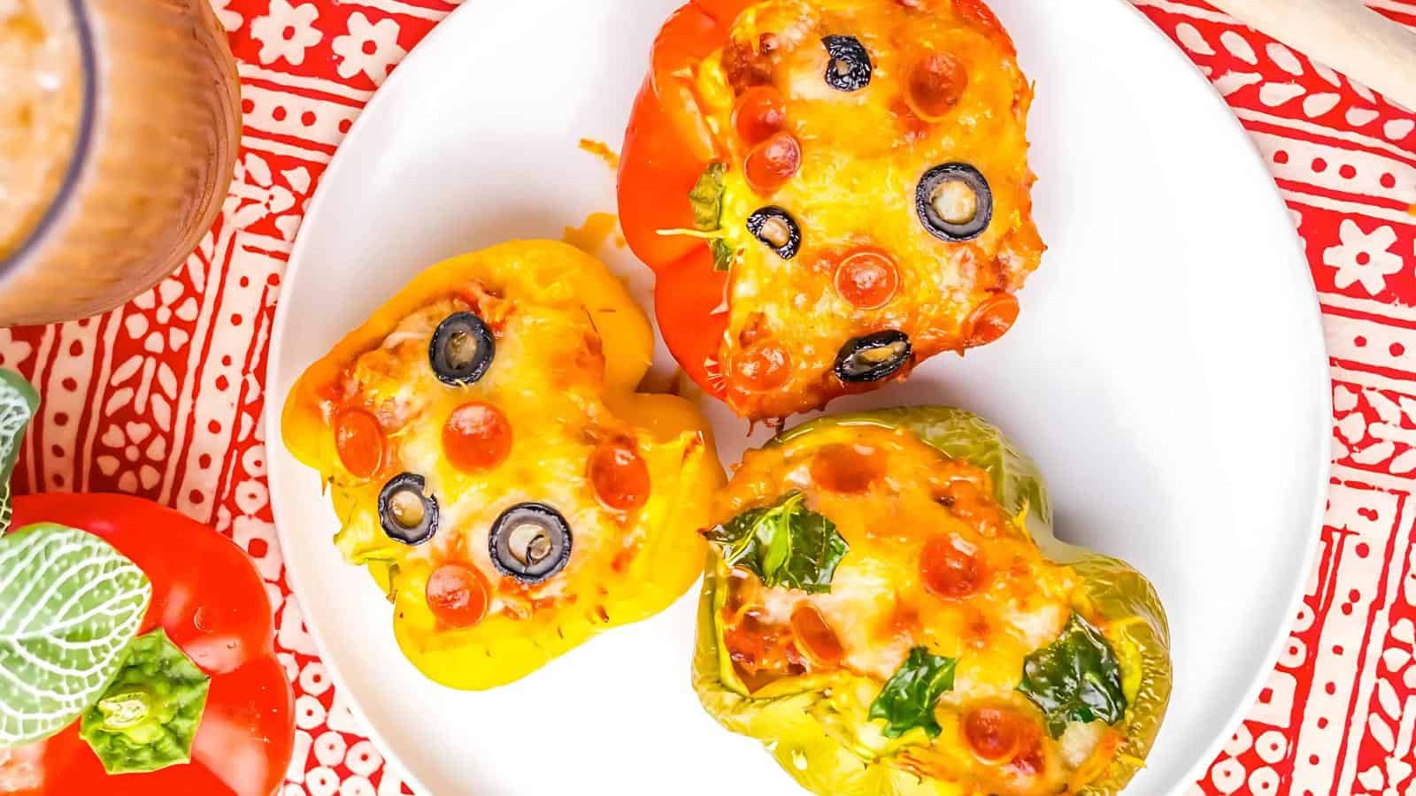 Three stuffed bell peppers topped with melted cheese, mini pepperoni, olives, and herbs on a white plate. A patterned red and white cloth and a leafy garnish are partially visible.
