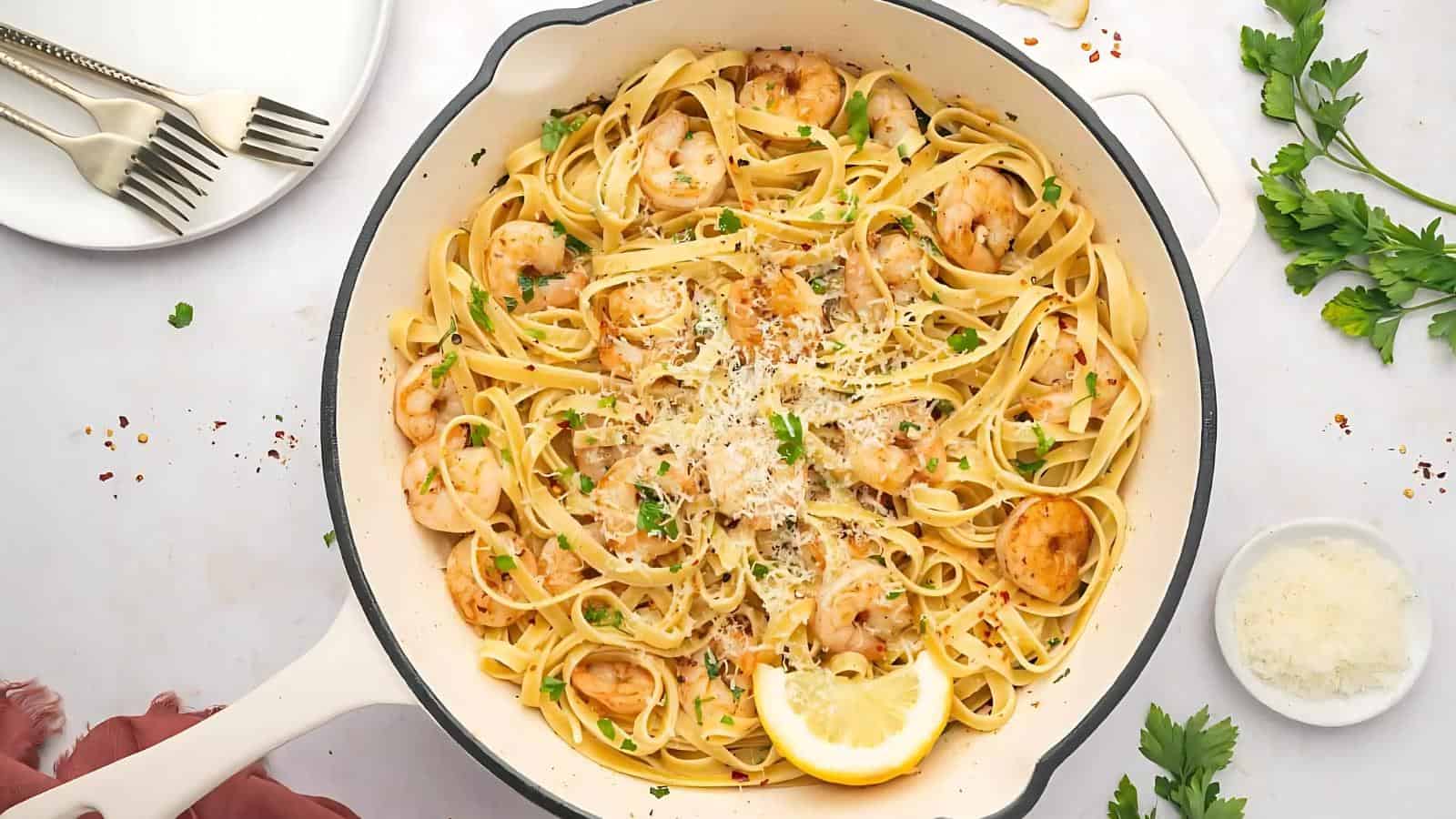 A pan of fettuccine with shrimp, garnished with grated cheese, lemon slice, and parsley. Plates, forks, parsley, and Parmesan are nearby.