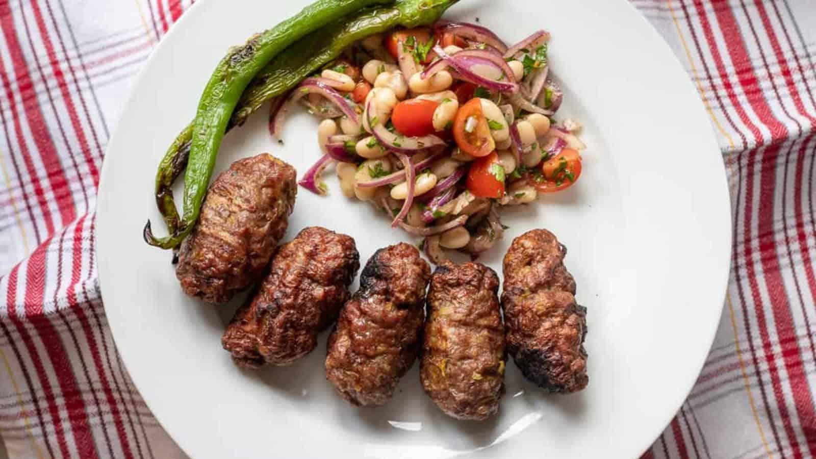 Plate with five grilled meat pieces, cooked green pepper, and a salad of beans, tomatoes, and red onions on a red and white checkered cloth.