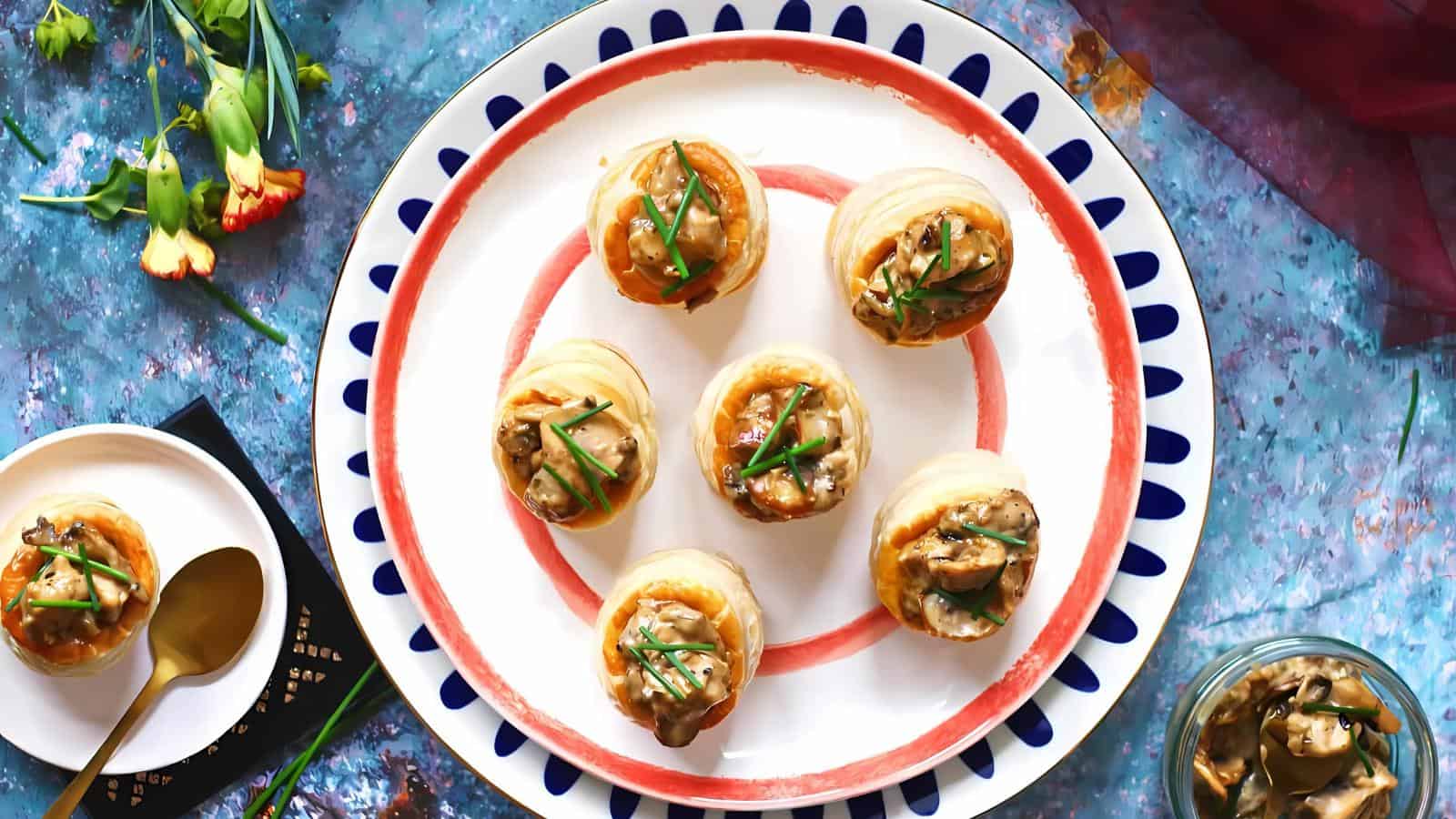 A plate with seven puff pastry appetizers filled with mushrooms and garnished with herbs. Surrounding the plate are flowers, utensils, and a jar with more filling.