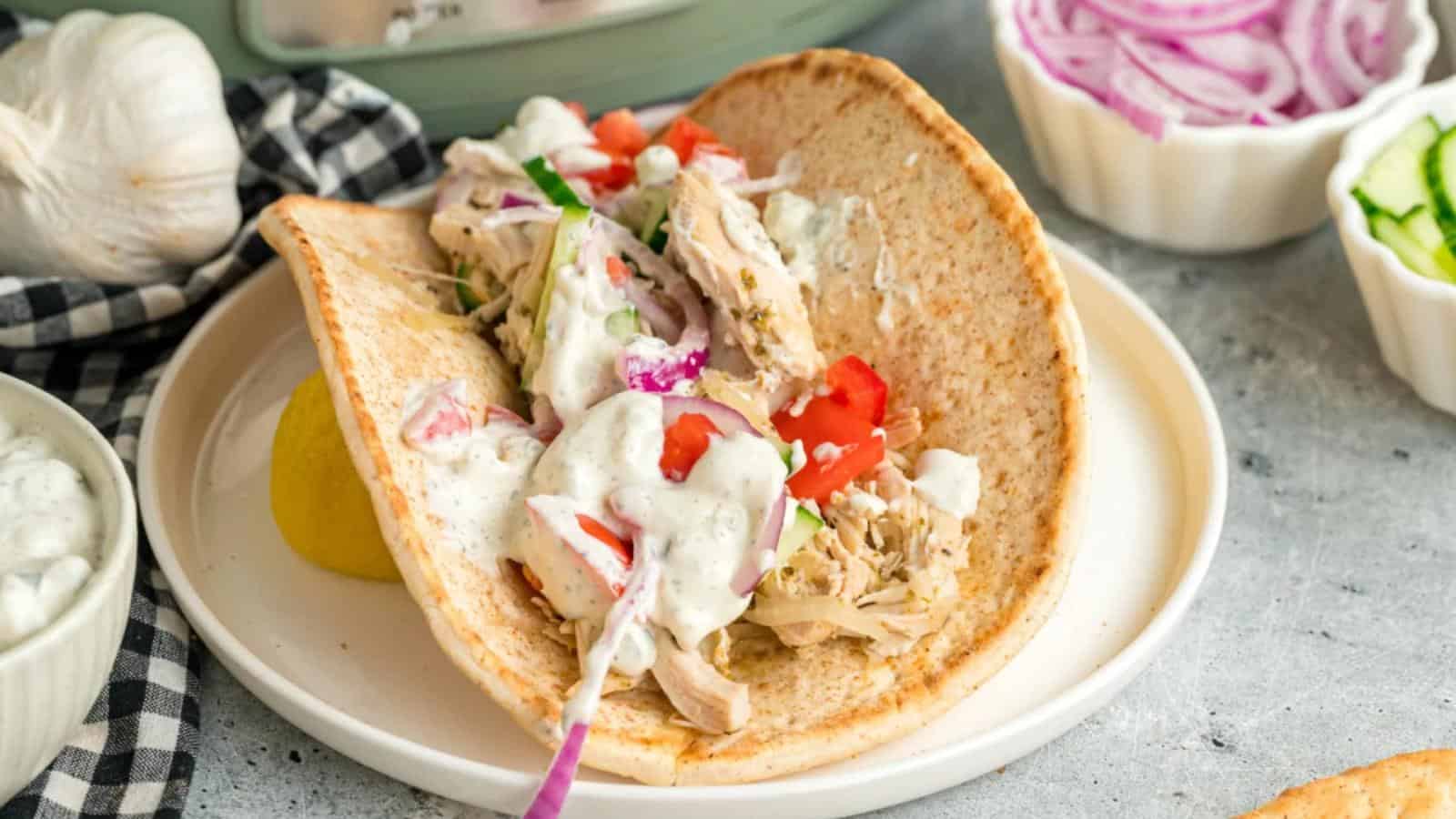 A pita filled with shredded chicken, diced tomatoes, onions, and a creamy white sauce, served on a white plate with a half lemon on the side.