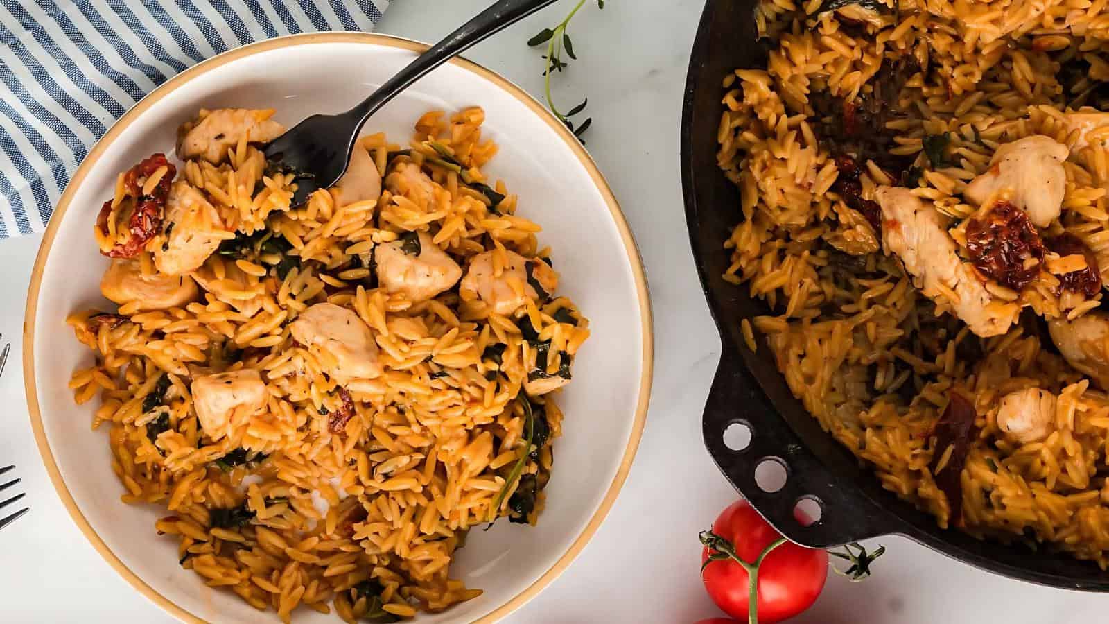 A bowl and a skillet filled with orzo pasta, chicken pieces, and sun-dried tomatoes. A fork rests in the bowl. Fresh tomatoes and a striped cloth are nearby.