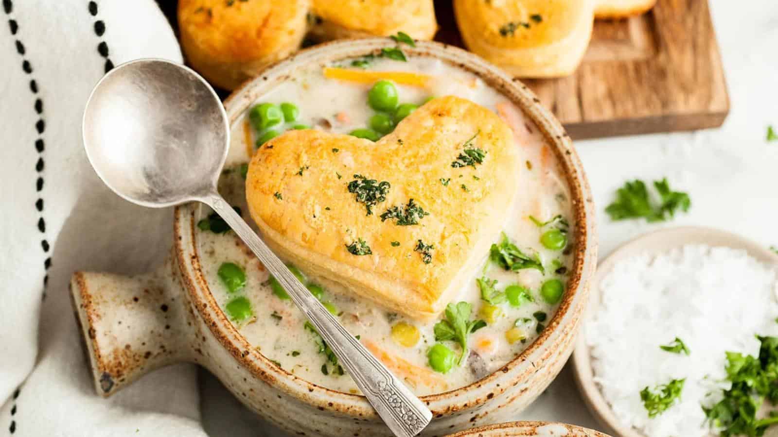 Heart-shaped pastry on a bowl of creamy soup with peas and herbs, accompanied by a spoon. Biscuits and a small dish of salt are in the background.