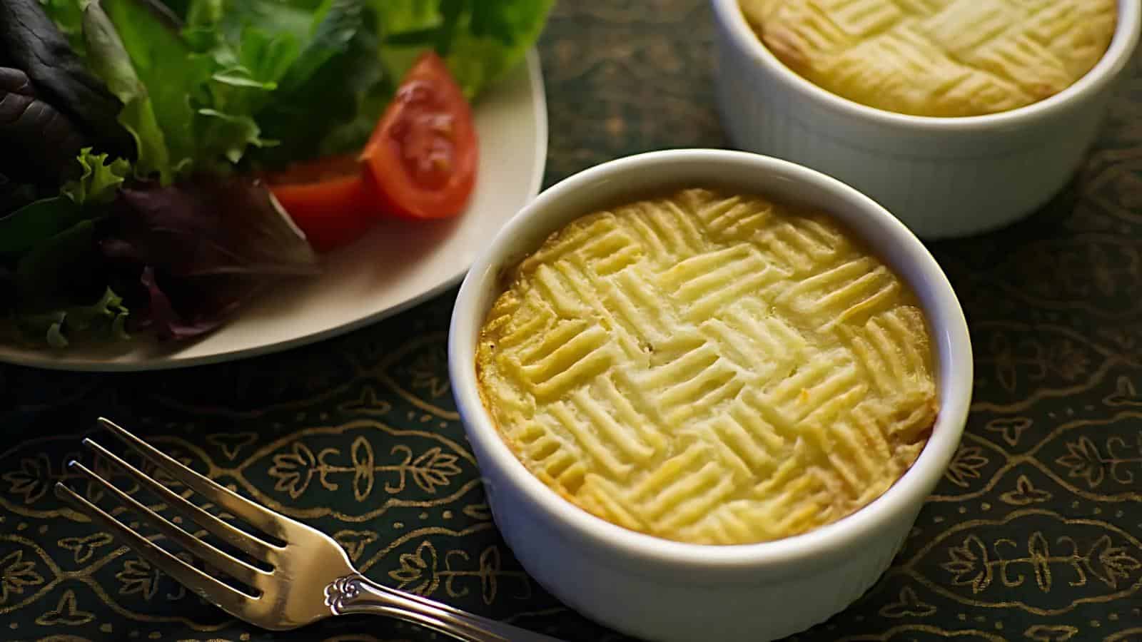 Two ramekins of baked shepherd's pie with a patterned top beside a plate of mixed greens and sliced tomato, and a silver fork on a decorative tablecloth.