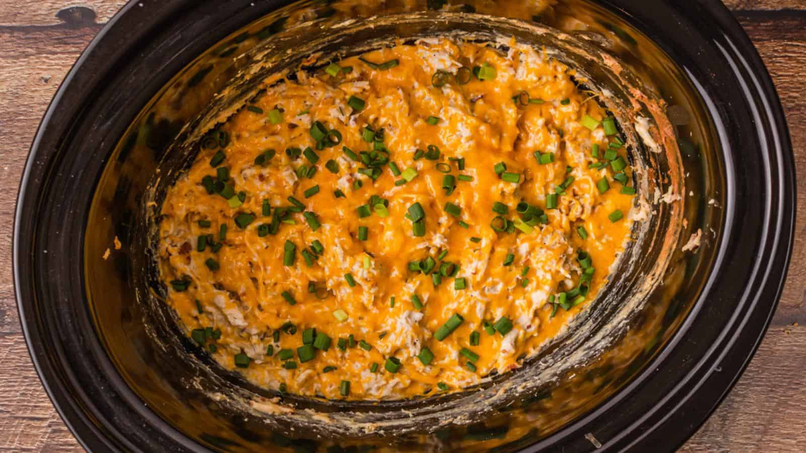 Cheesy dip in a slow cooker topped with chopped green onions on a wooden surface.