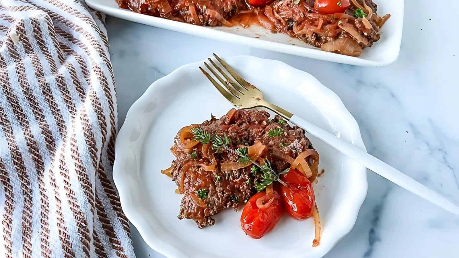 Plate of cooked liver with onions and cherry tomatoes garnished with herbs on a white plate. Fork beside the dish, with a striped cloth nearby.