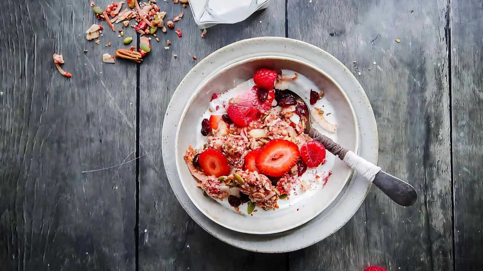A bowl of yogurt topped with granola, sliced strawberries, and raspberries on a rustic wooden table.