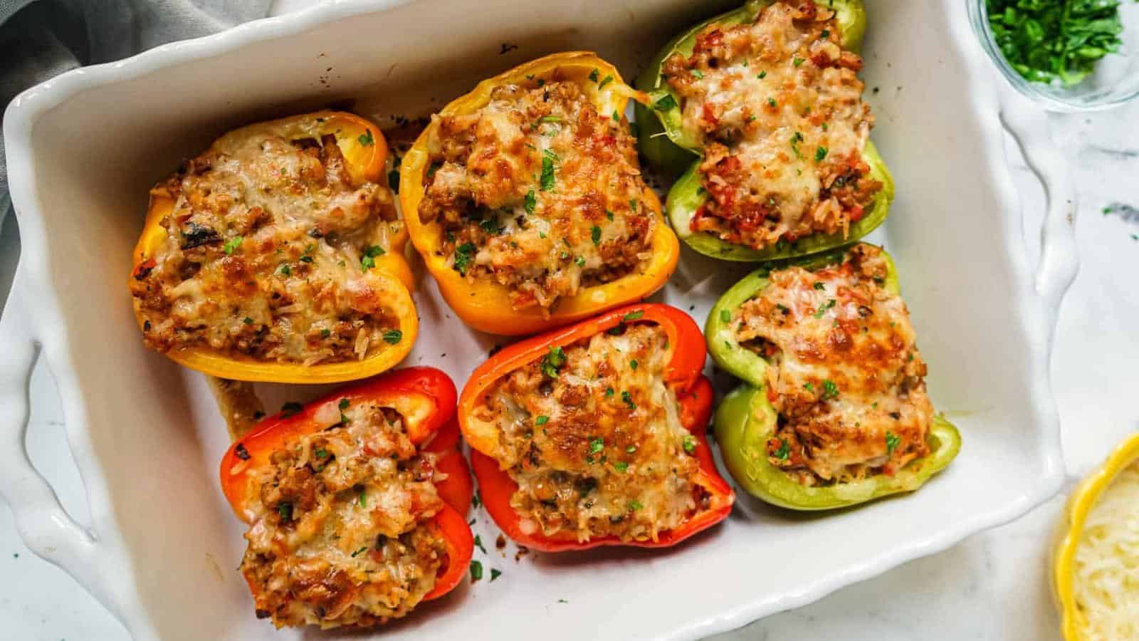 A white dish with six baked stuffed bell peppers in red, yellow, and green, filled with a seasoned mixture and topped with melted cheese.