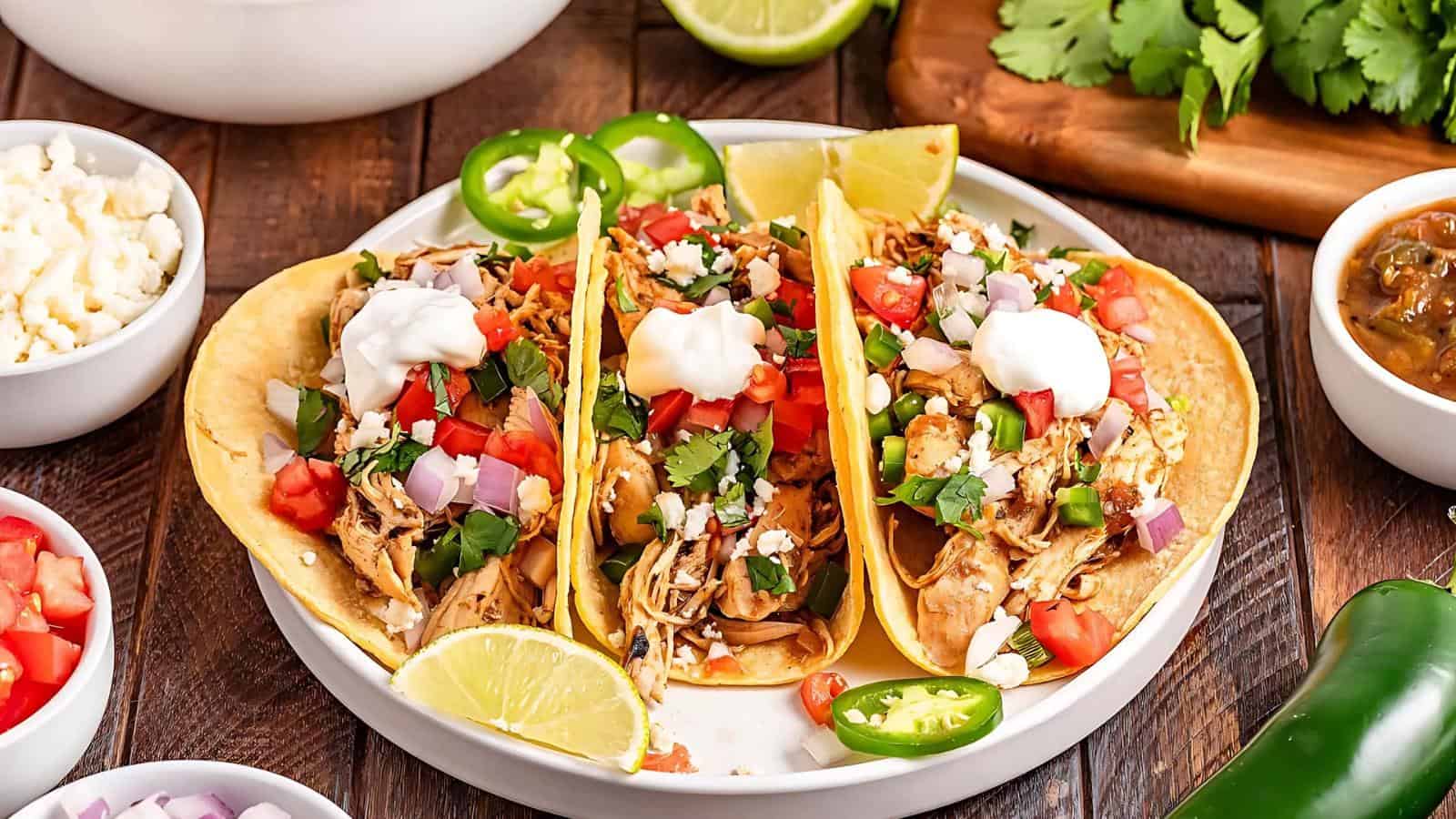 A plate of chicken tacos garnished with cilantro, diced tomatoes, onions, jalapeños, sour cream, and lime wedges, surrounded by bowls of ingredients.
