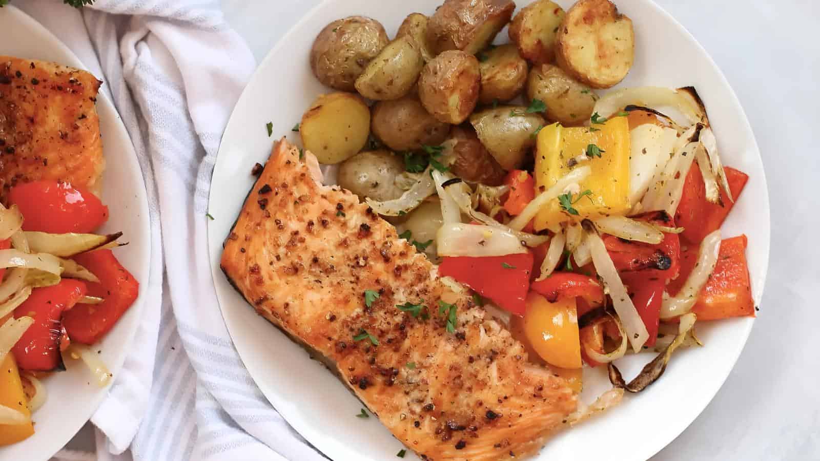 Plate of baked salmon with roasted potatoes, bell peppers, and onions, garnished with herbs, on a white plate near a striped towel.