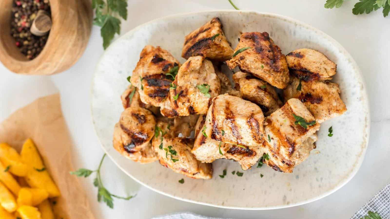 Grilled chicken pieces garnished with herbs on a white oval plate. A wooden bowl with peppercorns and sliced yellow vegetables are nearby.