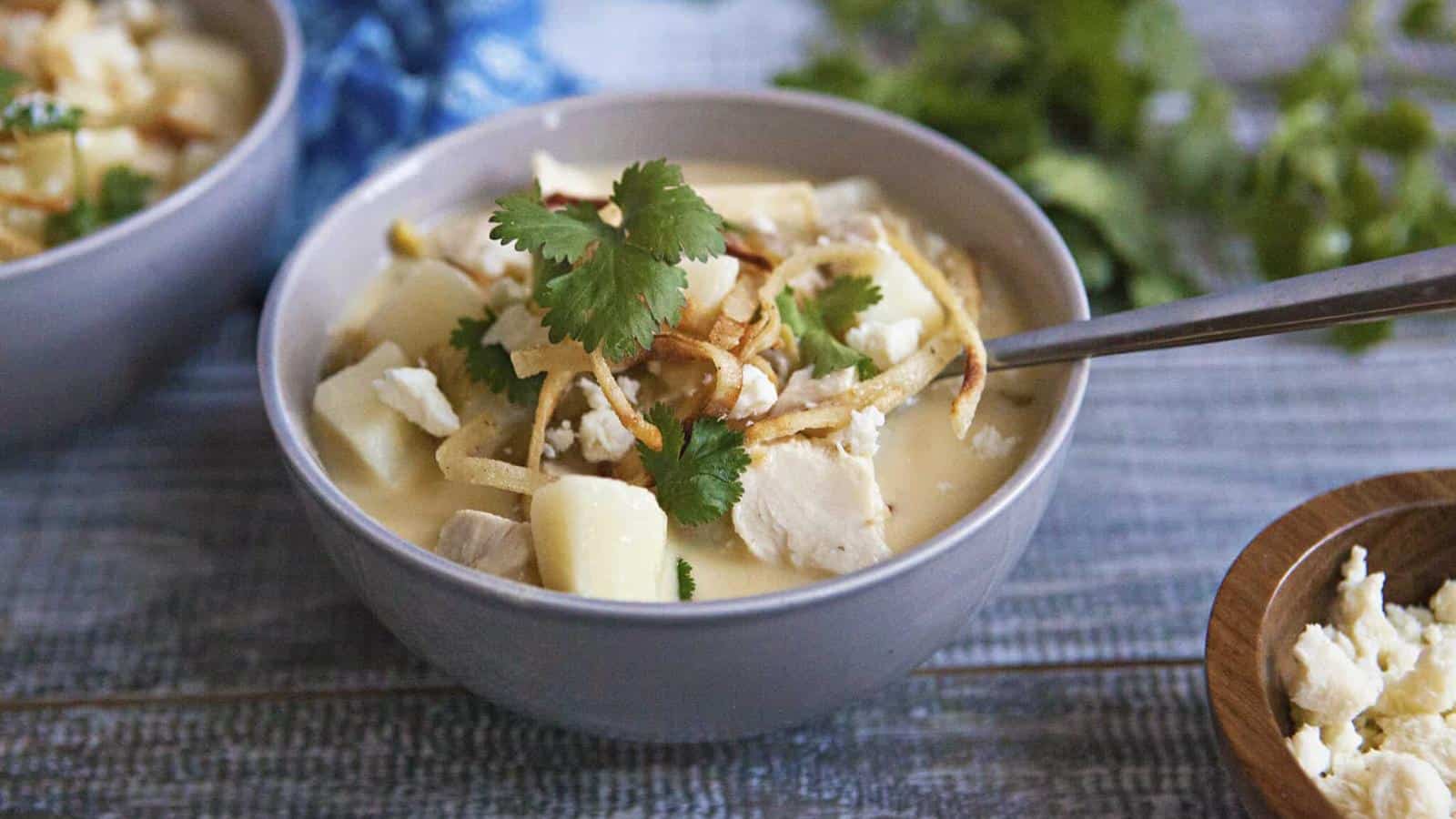 A bowl of creamy soup with chunks of chicken, potatoes, garnished with cilantro and crispy toppings. A spoon rests inside the bowl.