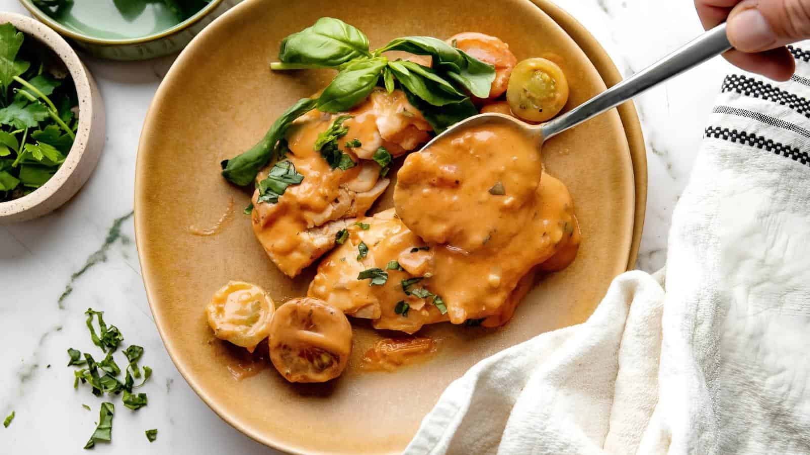 A plate with chicken in tomato cream sauce, garnished with basil leaves. A spoon is lifting sauce from the dish. Sliced tomatoes are on the side. A cloth napkin partially covers the plate.
