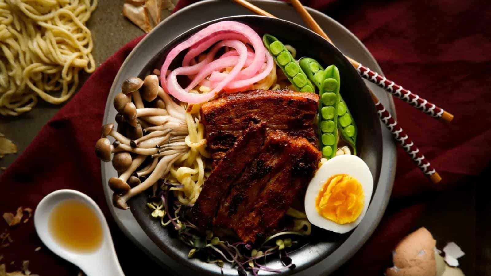A bowl of ramen with grilled meat, boiled egg, mushrooms, pickled onions, snap peas, and noodles. Chopsticks rest on the bowl's edge, and a small dish of sauce is nearby.