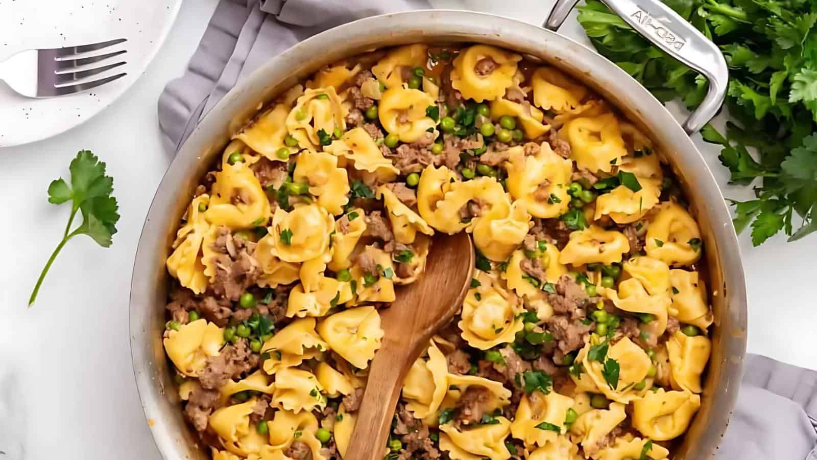 Skillet filled with tortellini pasta, ground meat, and peas, garnished with parsley. A wooden spoon is in the pan. A sprig of parsley and a fork are nearby on a gray cloth.