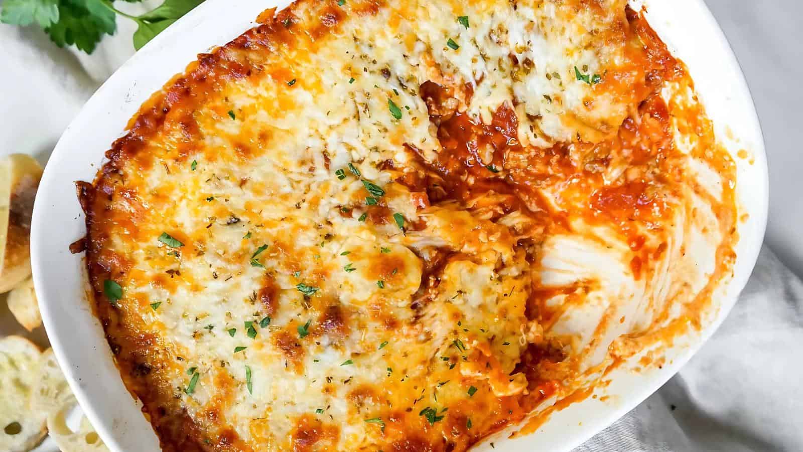 Baked lasagna in a white casserole dish, partially served, with a golden-brown cheese crust and garnished with herbs.