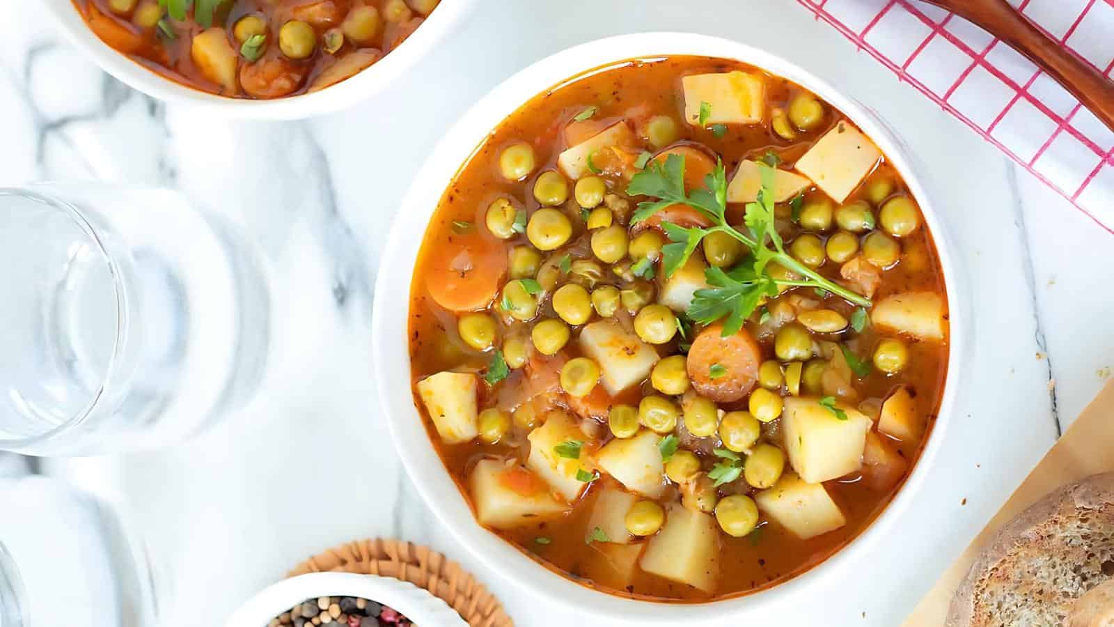 A bowl of vegetable stew with peas, potatoes, and carrots garnished with parsley, alongside a glass of water and a bowl of pepper.