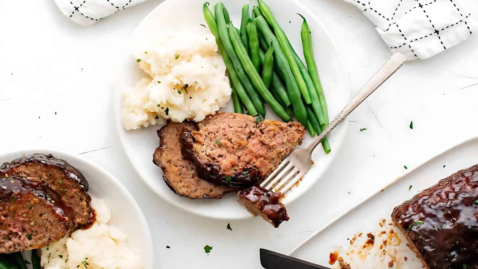 Plate of meatloaf with gravy, mashed potatoes, and green beans, accompanied by a fork.