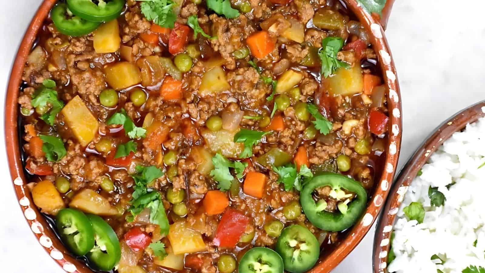 A bowl of picadillo with ground meat, potatoes, carrots, peas, tomatoes, cilantro, and sliced jalapeños, next to a side of white rice.