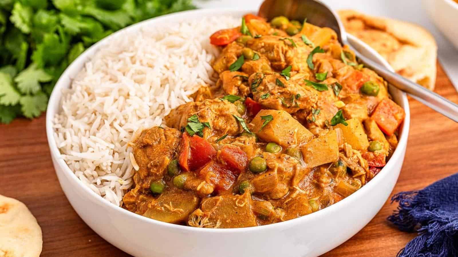 A bowl with white rice and a curry dish containing chicken, peas, carrots, and potatoes, garnished with cilantro. A spoon is placed in the bowl, with naan bread on the side.