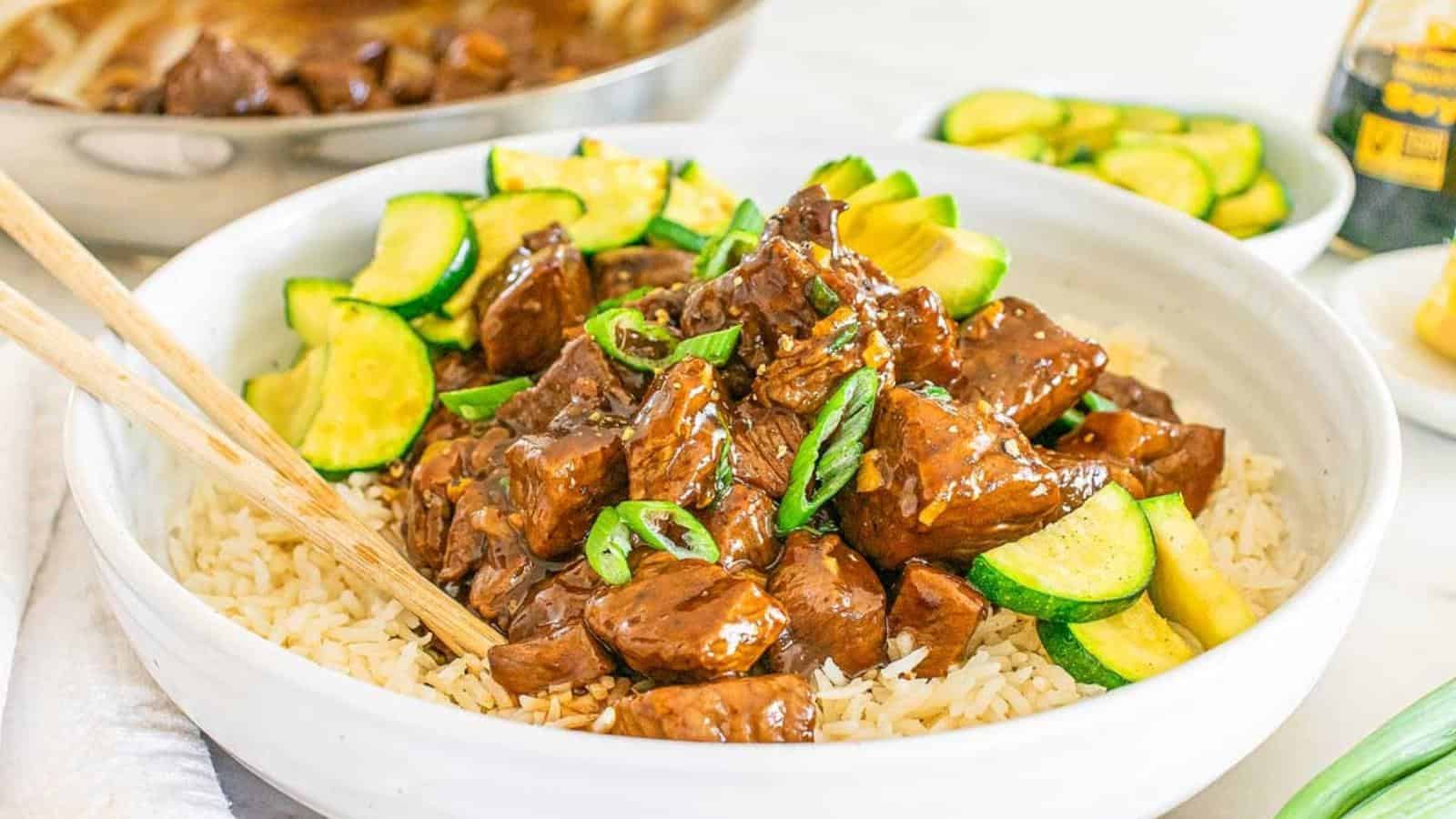A bowl of rice topped with glazed beef chunks, sliced zucchini, and chopped green onions, with chopsticks resting on the side.