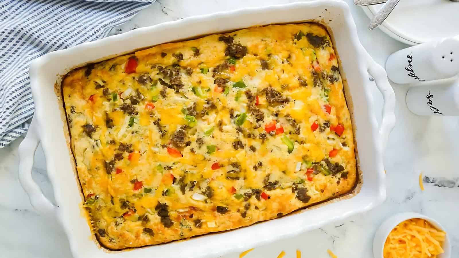 A baked breakfast casserole in a rectangular dish with visible ground meat, cheese, and diced vegetables. A striped cloth, utensils, and a small bowl of shredded cheese are nearby.