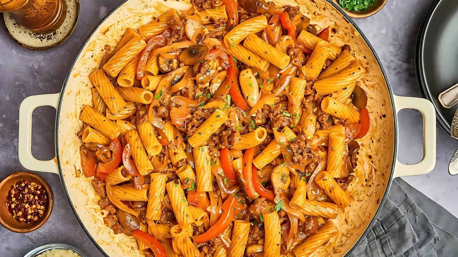 A pot of rigatoni pasta with red bell peppers, mushrooms, and ground meat in a seasoned sauce on a gray tabletop.