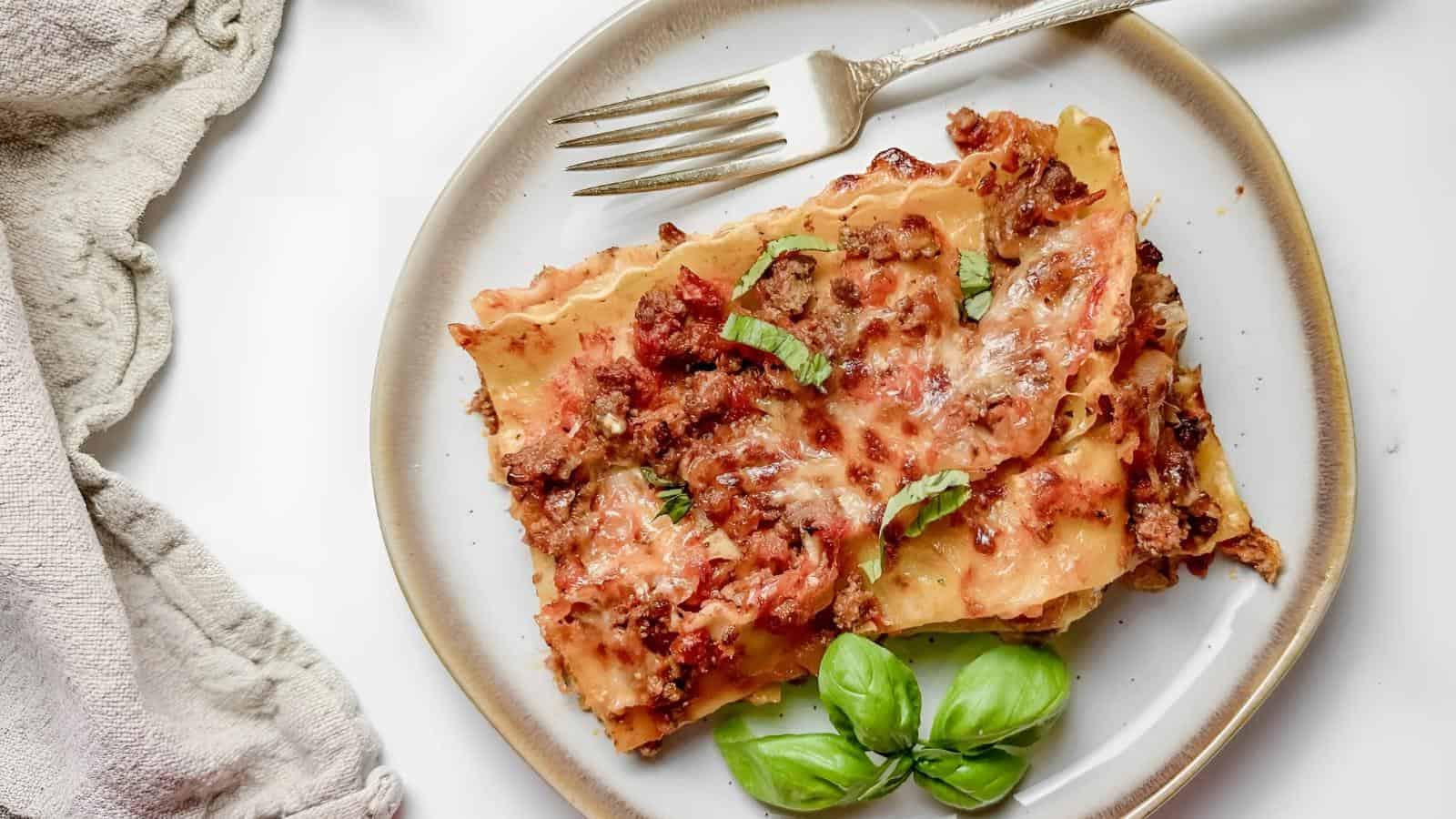 A slice of lasagna on a white plate, garnished with fresh basil leaves. A fork rests beside it, next to a beige napkin.