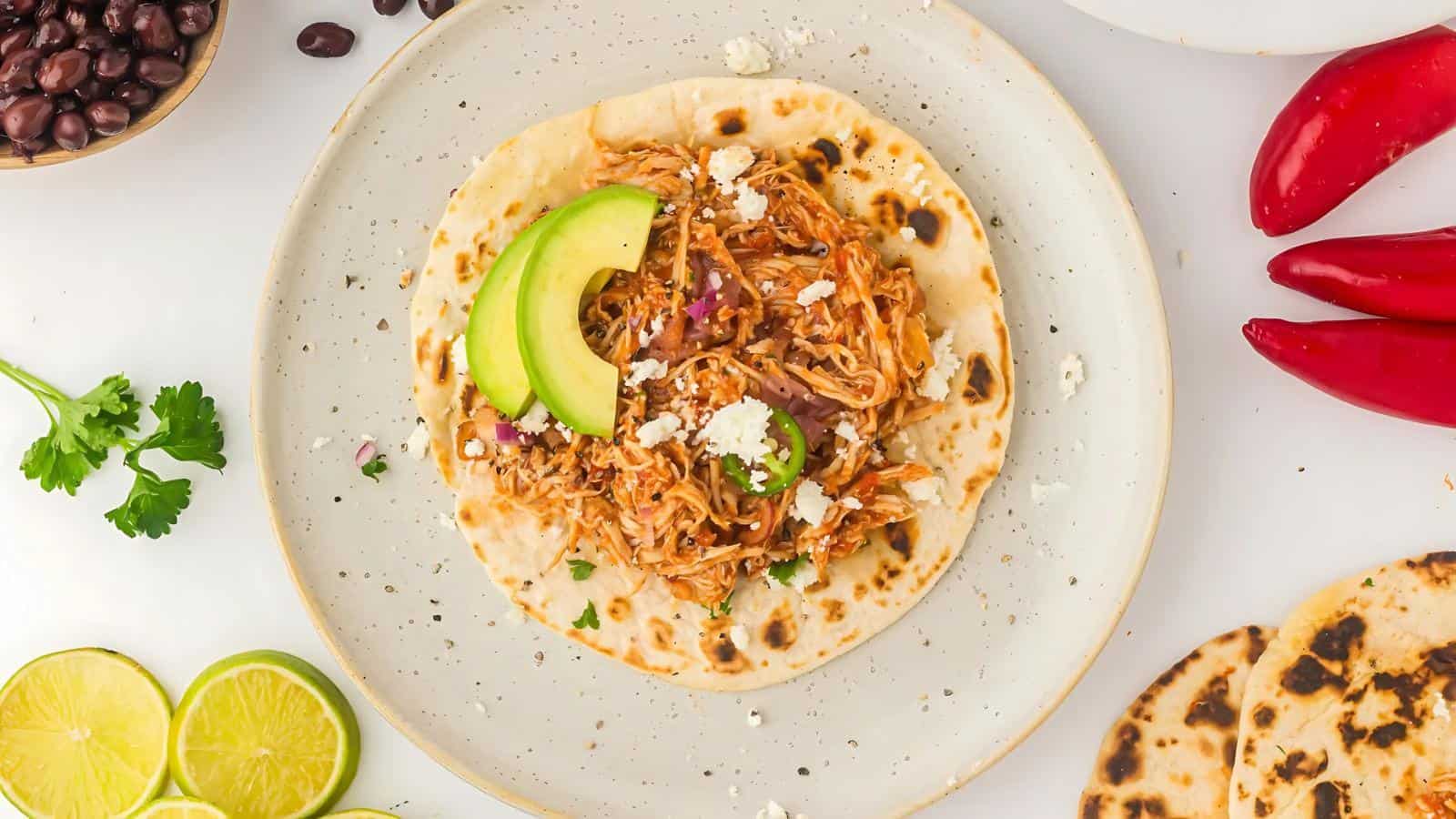 A tortilla topped with shredded chicken, avocado slices, and crumbled cheese on a plate. Nearby are lime halves, red peppers, cilantro, black beans, and extra tortillas.