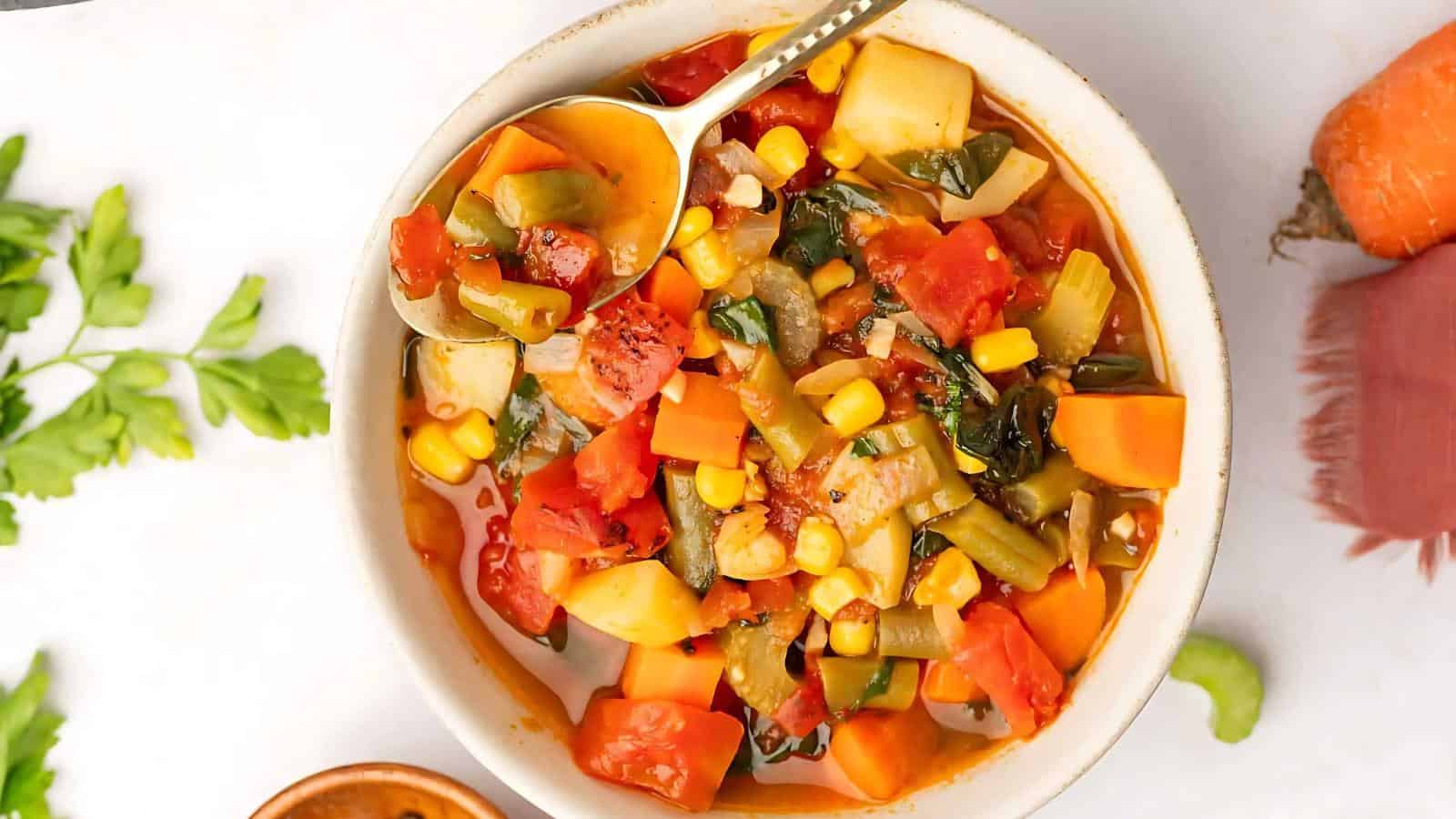 A bowl of vegetable soup with visible corn, tomatoes, carrots, green beans, and spinach. A spoon rests inside the bowl. Parsley and a carrot are beside the bowl on the table.