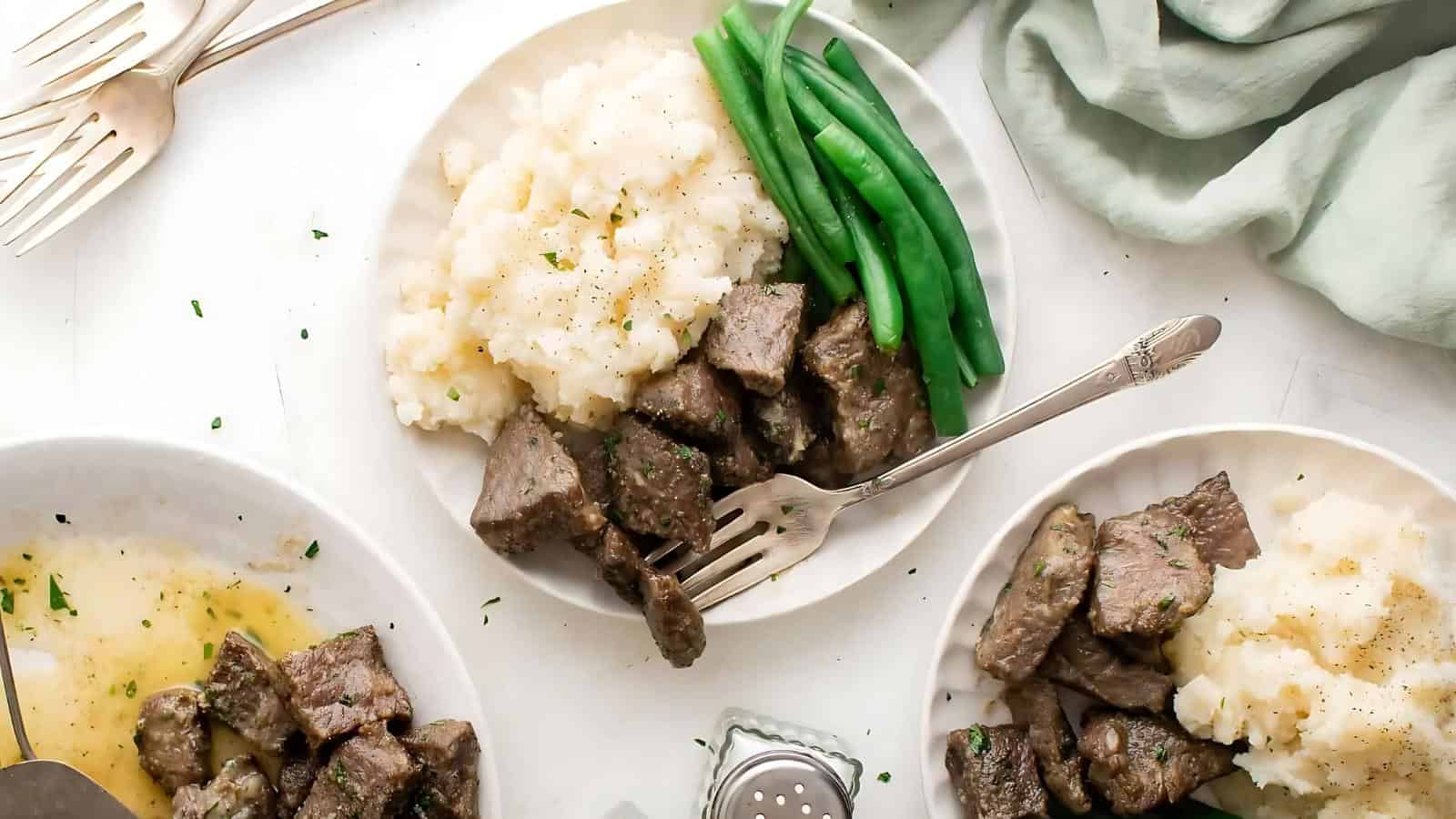 Plates of diced beef with mashed potatoes and green beans, served on a light background with forks and a salt shaker nearby.