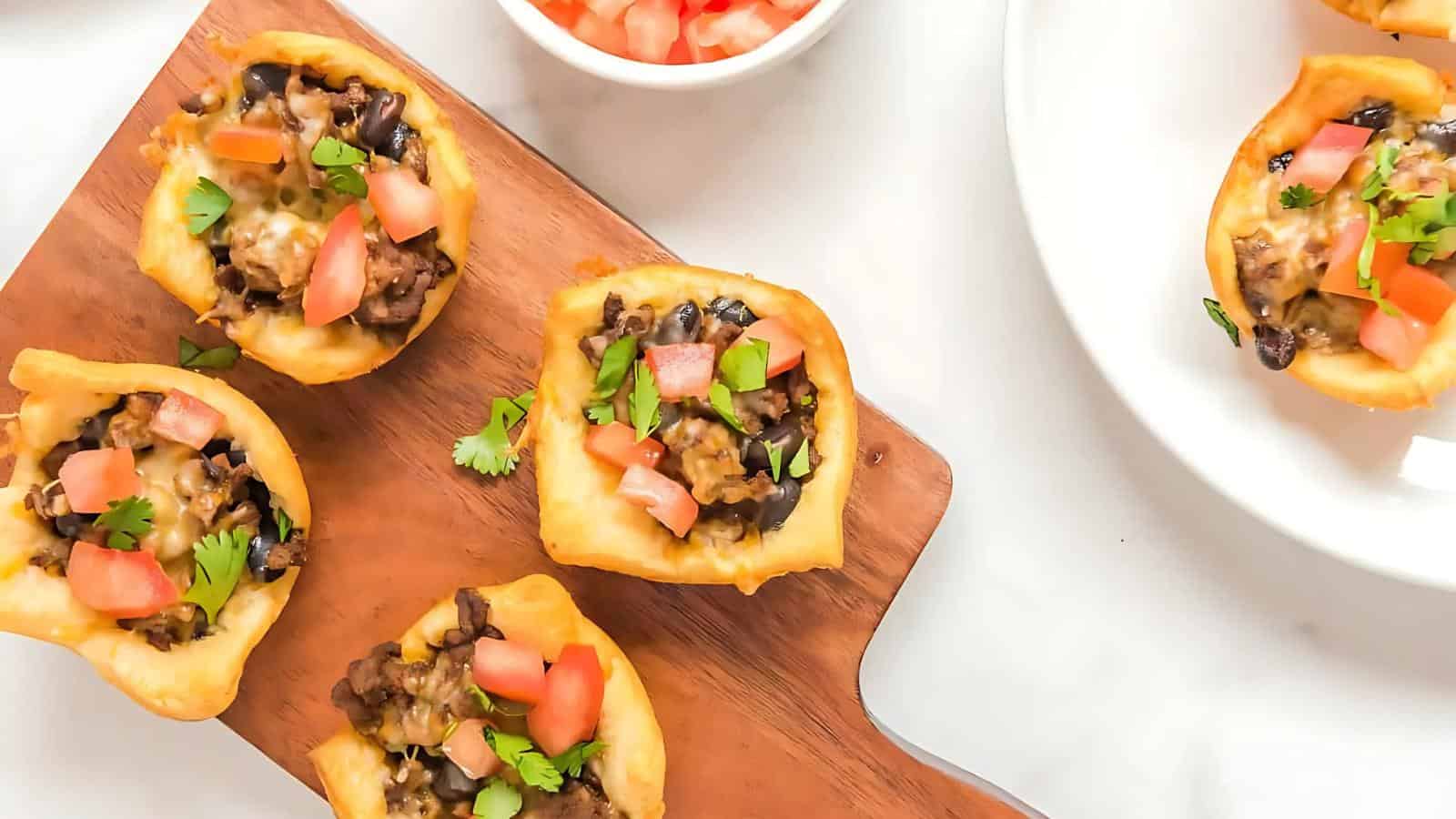 Mini taco cups on a wooden board, filled with beef, black beans, cheese, tomatoes, and cilantro. A bowl of diced tomatoes is nearby.