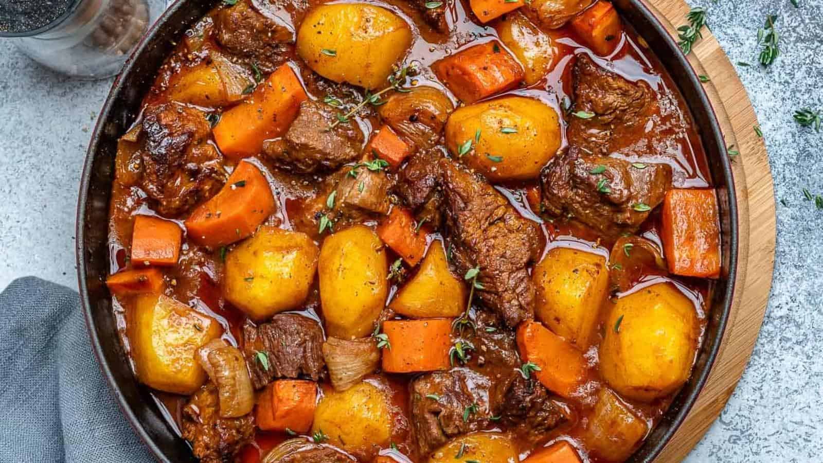 A hearty beef stew with chunks of beef, potatoes, and carrots in a rich brown sauce, garnished with herbs, served in a black bowl on a wooden board.